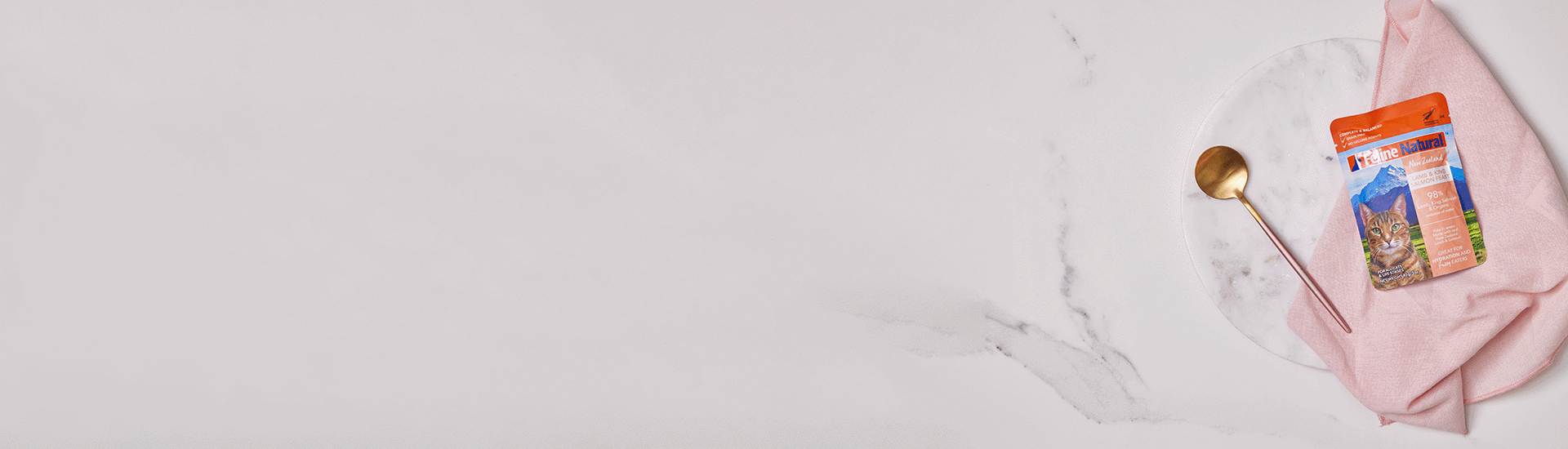 Feline Natural Lamb & King Salmon Pouch resting on a pink hand towel. A serving spoon is next to it